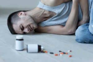 young man lying on the floor holding hands stomach while bottles with pills laying near him