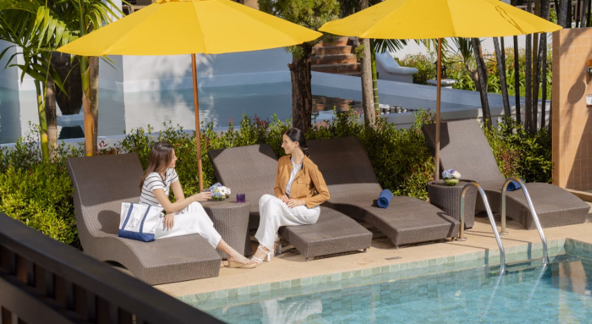 Two women relaxing and chatting on poolside lounge chairs under yellow umbrellas.