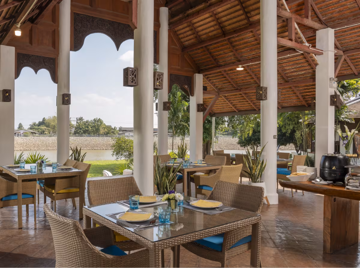 Open-air restaurant with set wicker dining tables under a teak roof overlooking the river at The Dawn
