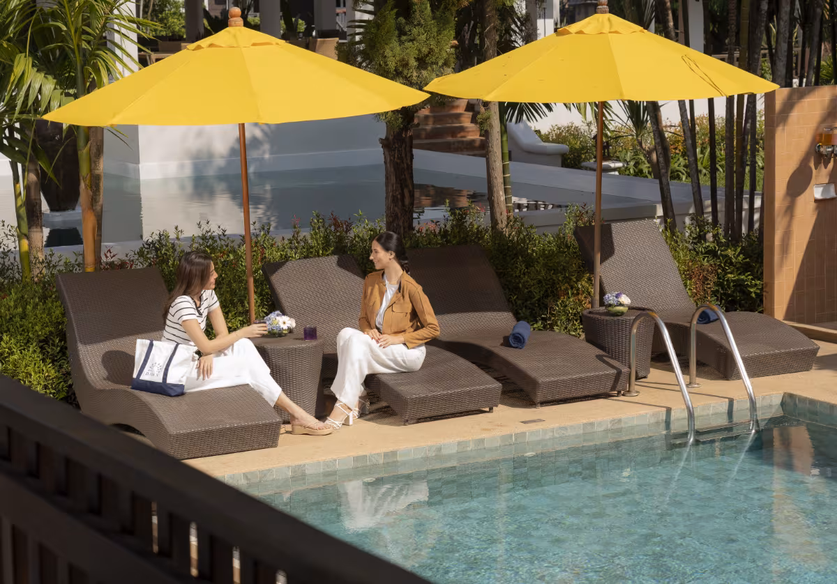 Two women chatting on loungers beneath yellow umbrellas beside the pool at The Dawn Rehab Thailand